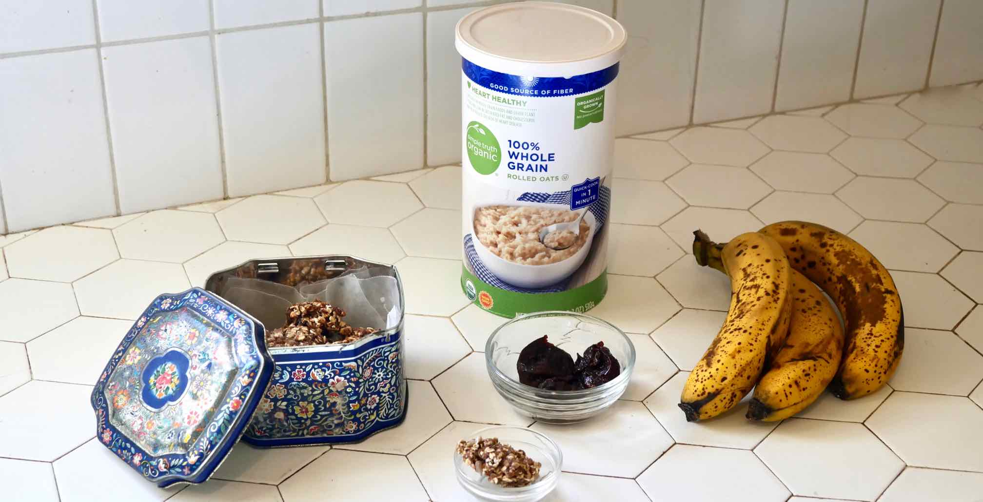 Cookie tin with healthy cookies, container of rolled oats, three prunes in a dish, one healthy cookie and three very ripe bananas