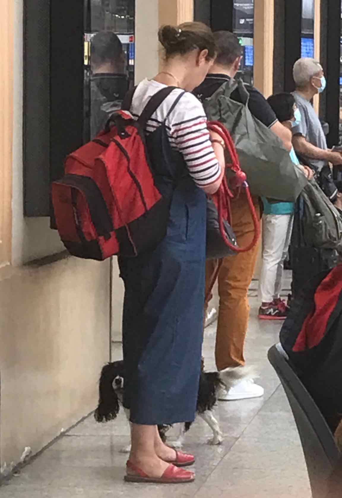 young woman wearing dungarees and marinière top with spaniel, red leash and red rucksack waiting for train