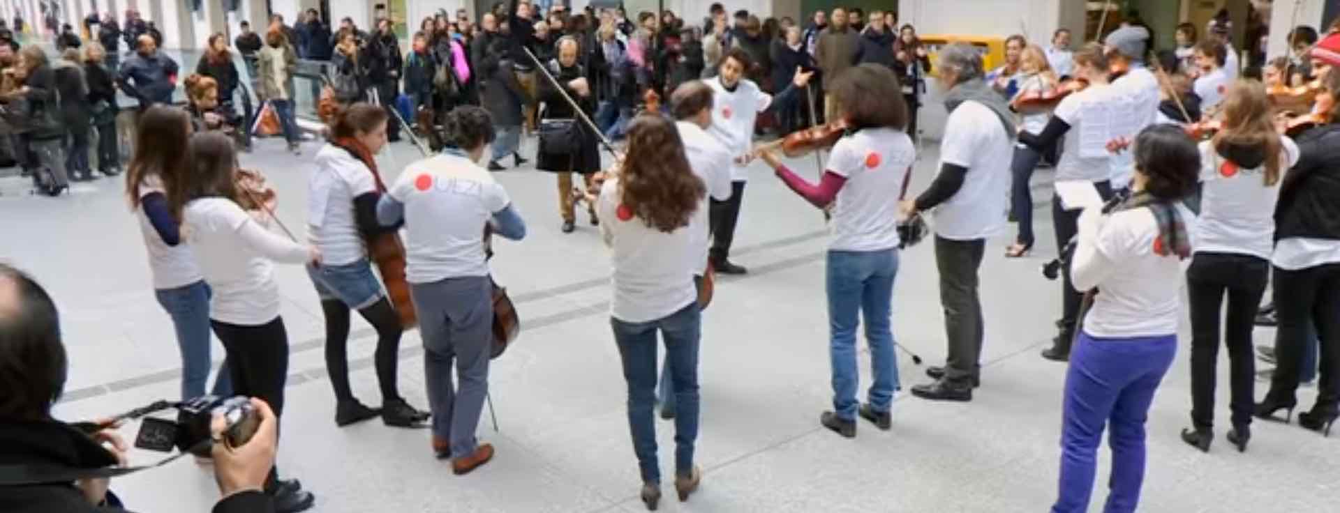 orchestra members with instruments perform at the Gare Saint-Lazare wearing jeans and white tee shirts with crowd watching