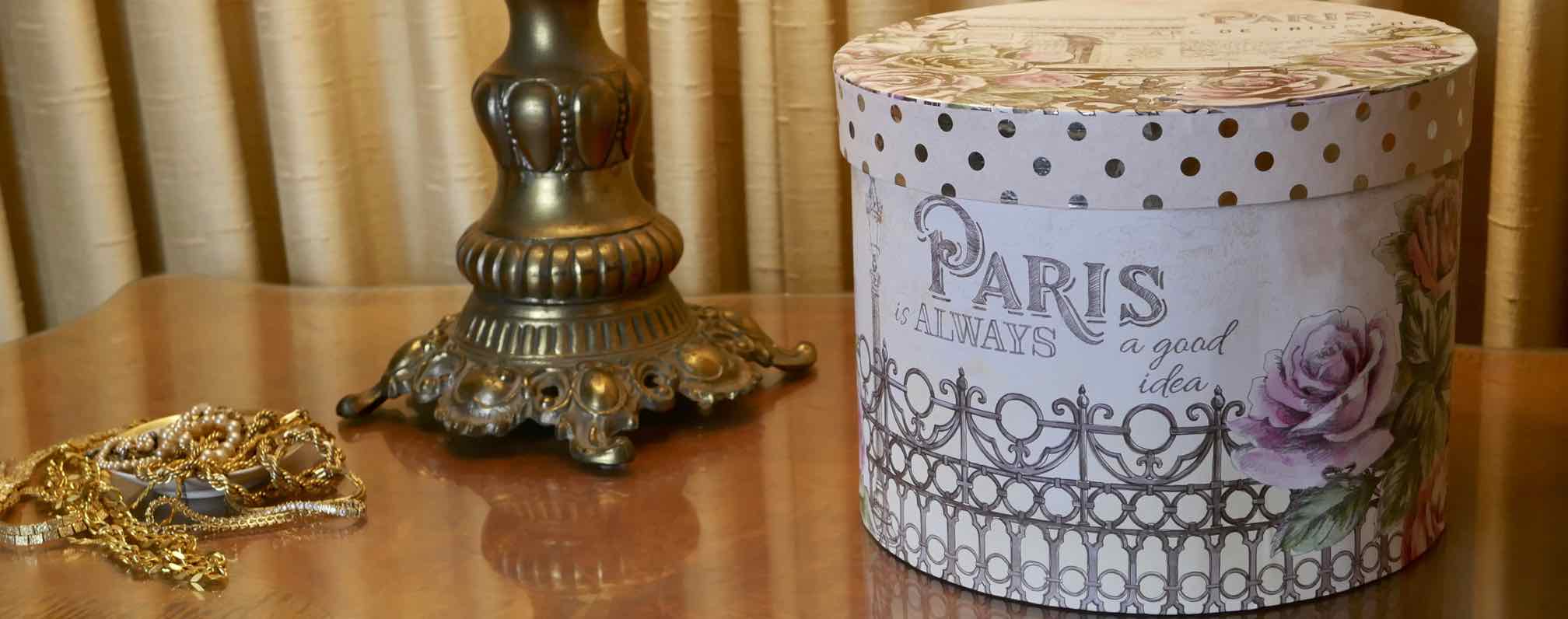 hat box with roses and inspription: Paris is always a good idea on table with lamp and assorted jewelry