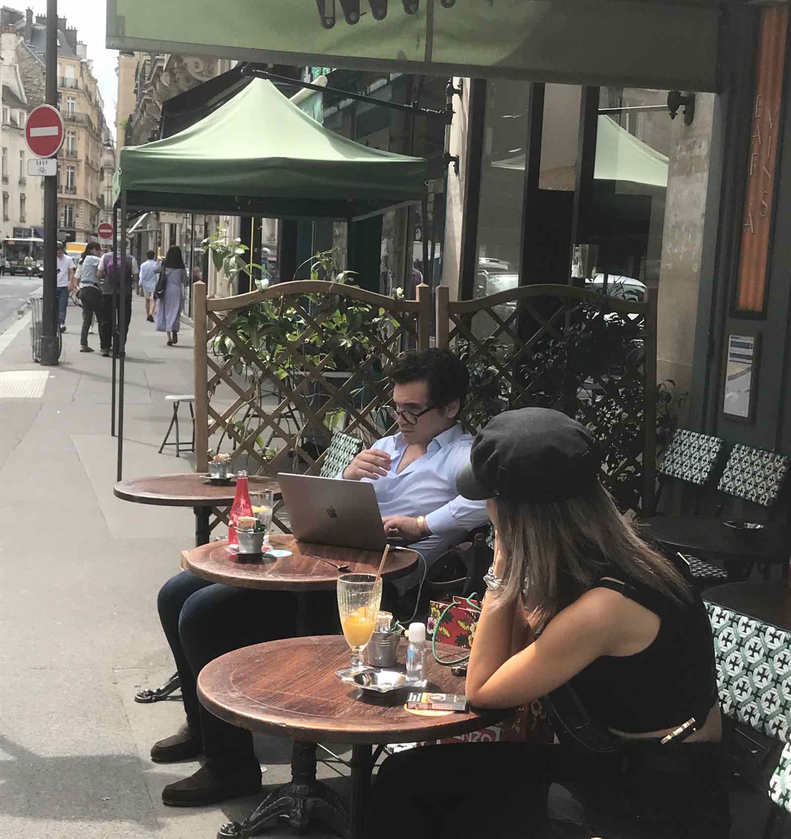 young woman on terrace wearing black butcher's boy cap, crop top and capri pants observing young man at next table writing novel (?) on his MacBook Pro