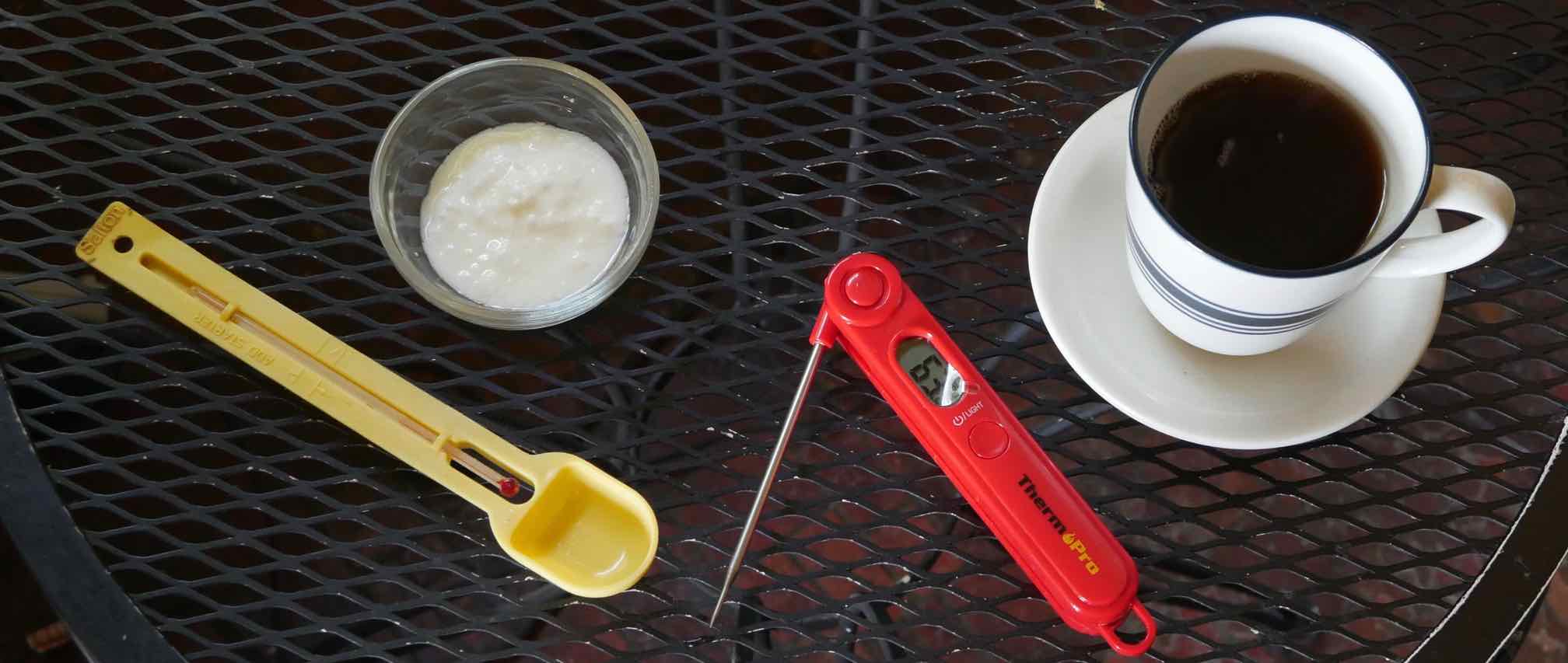 a measuring spoon themometer, a small dish of yougurt starter, a digital thermometer and a cup of hot tea in a blue and white mug.